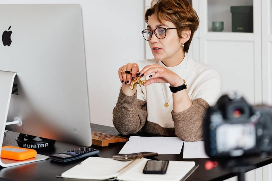 A professional woman engaged in a virtual meeting setup at her home desk, using a computer and camera.