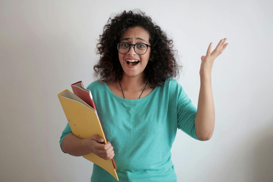 Dissatisfied annoyed woman with mouth opened wearing glasses and turquoise blouse looking away and screaming while standing against white wall with folders of documents and having problems in work